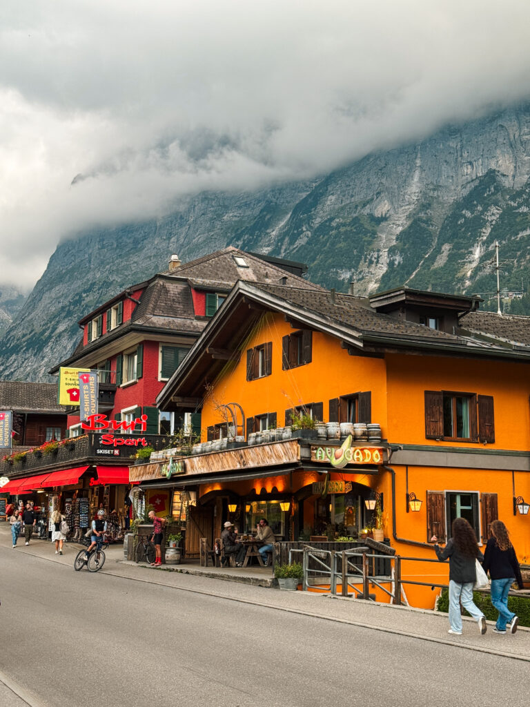 Street view in Grindelwald with colorful buildings, outdoor cafés, and shops set against dramatic alpine cliffs partly covered in clouds.