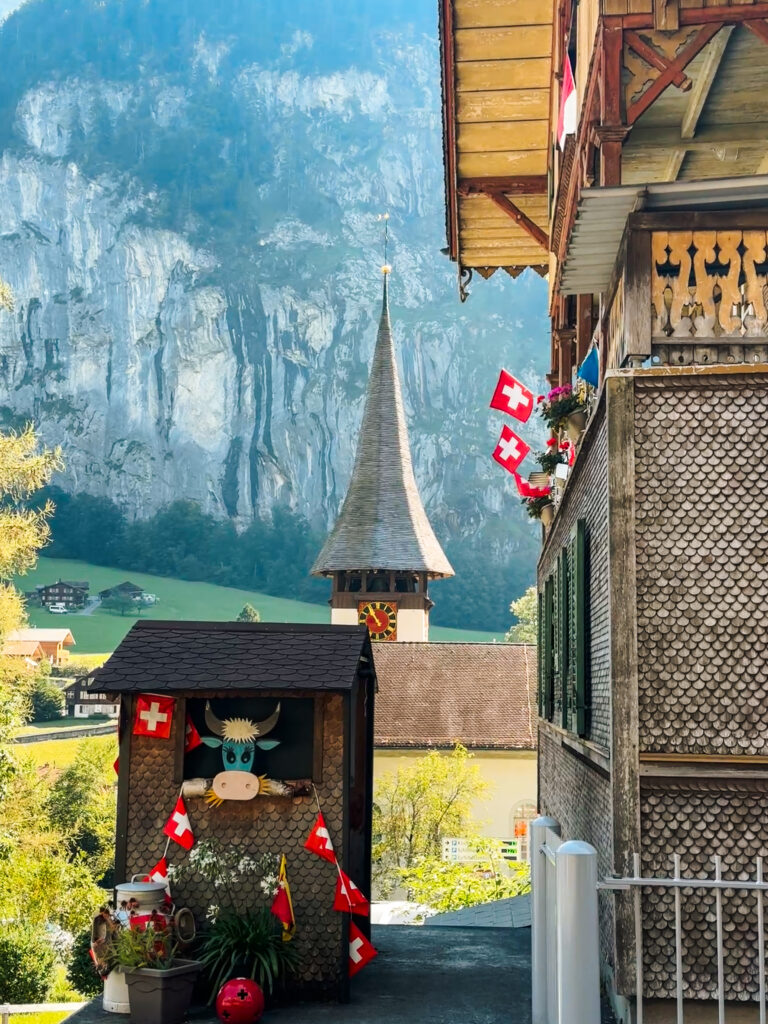 View of Lauterbrunnen church steeple framed by alpine houses and Swiss flags, with dramatic cliffs rising in the background.