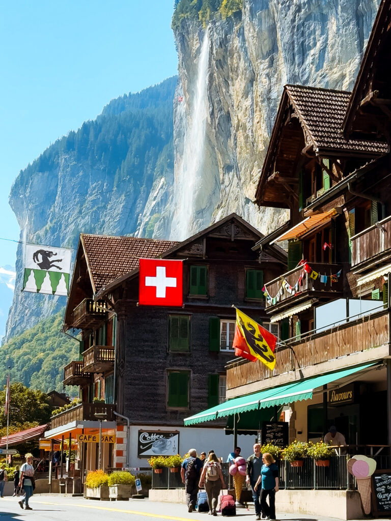 Street scene in Lauterbrunnen village with traditional Swiss chalets, flags, and Staubbach Falls cascading down the cliffside behind.