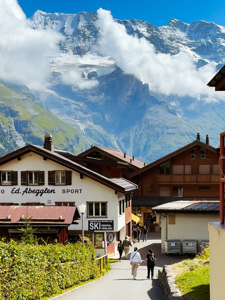 Street in Mürren lined with traditional alpine buildings and ski shops, with snow-capped Swiss Alps and dramatic cliffs rising behind.