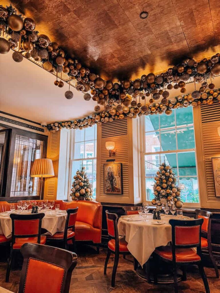 A cozy corner of a London restaurant 34 Mayfair during Christmas season with orange leather chairs, white table settings, and small decorated Christmas trees by the windows under golden ceiling garlands.