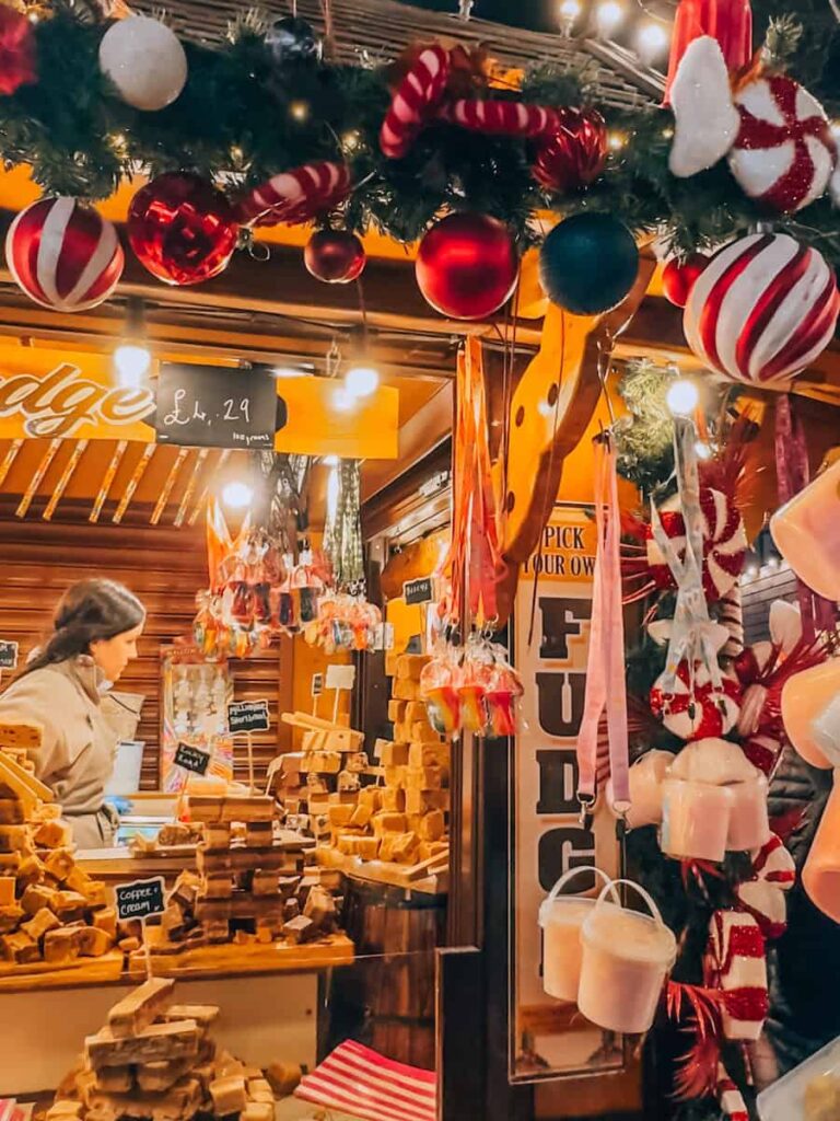 A festive Christmas market stall selling homemade fudge, decorated with garlands, candy canes, and ornaments, as a vendor arranges stacks of fudge under the warm glow of holiday lights during Christmas in London. 