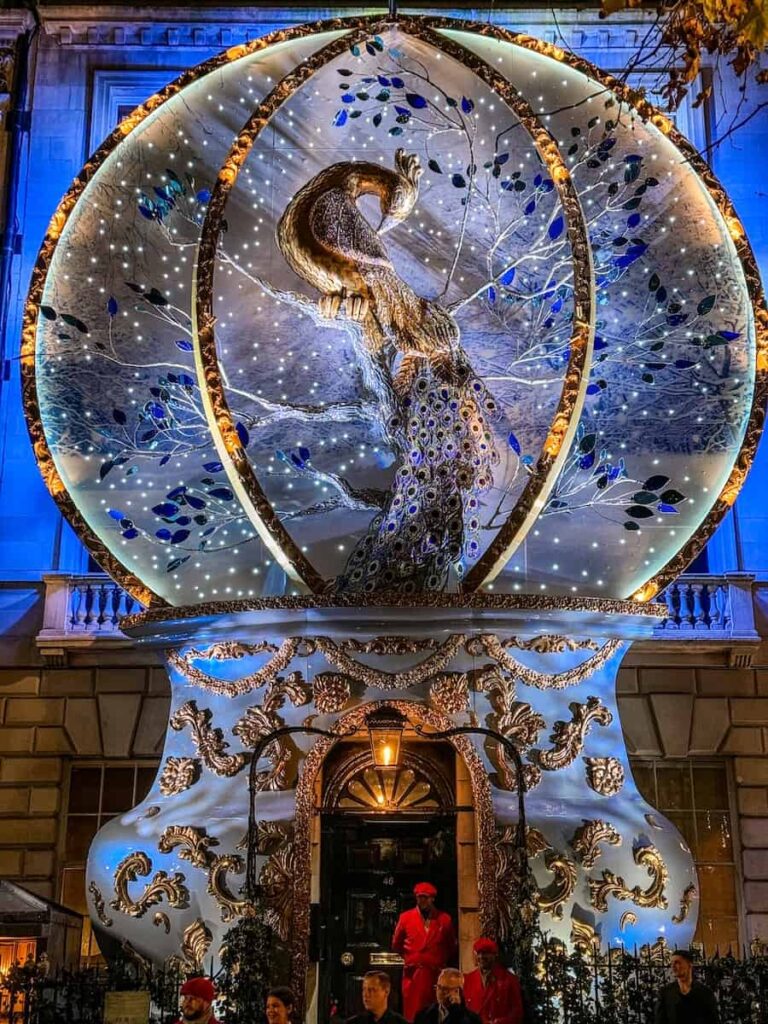 The exterior of a London building at Annabel's adorned with an elaborate holiday display featuring a majestic golden peacock perched inside a giant illuminated ornament decorated with sparkling blue and silver details.
