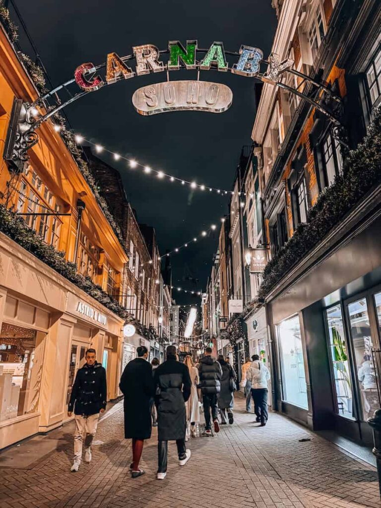 Carnaby Street in London at night, decorated with string lights and a colorful illuminated “Carnaby Soho” sign above the bustling pedestrian walkway filled with shoppers.