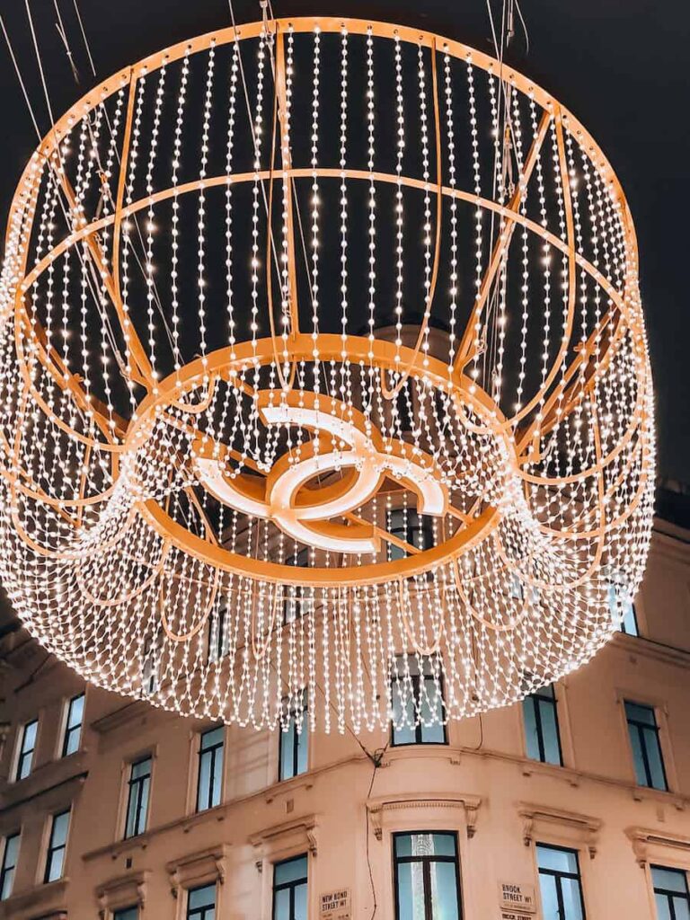 A large illuminated Chanel logo chandelier made of cascading strings of white lights, glowing against the backdrop of a historic London building at night.