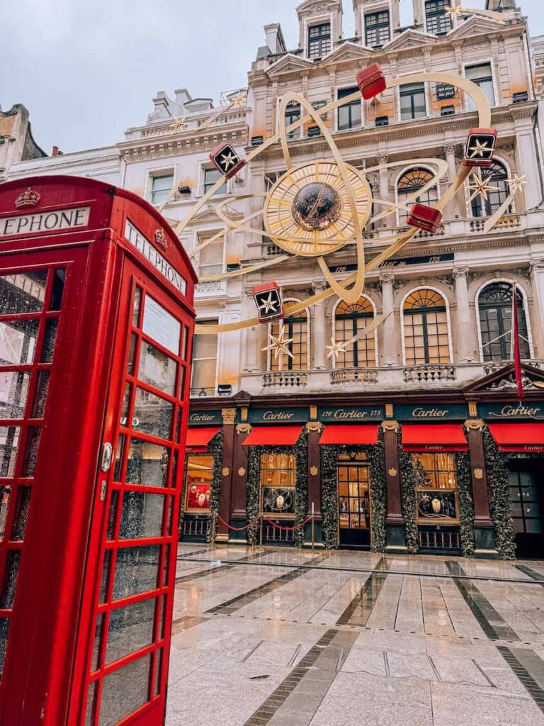 A festive London street scene featuring a classic red telephone booth beside the Cartier store on New Bond Street, beautifully decorated for Christmas with red and gold ornaments, garlands, and a large golden clock display above the storefront.