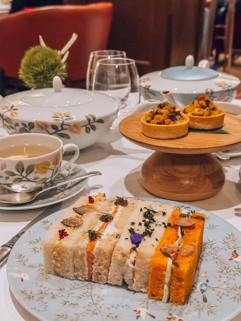 A beautifully arranged afternoon tea setting featuring assorted finger sandwiches with elegant garnishes, mini savory tarts, floral teacups, and teapots with botanical patterns on a white tablecloth.