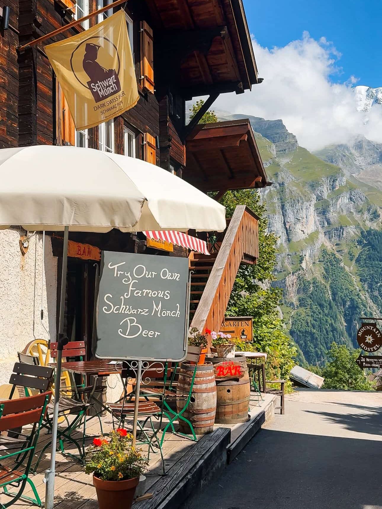 A charming mountain café in Gimmelwald, Switzerland, with wooden chalet architecture, flower pots, and a sign inviting visitors to try Schwarz Mönch beer. The terrace has colorful chairs under a white umbrella, and the backdrop features dramatic alpine cliffs and blue skies.