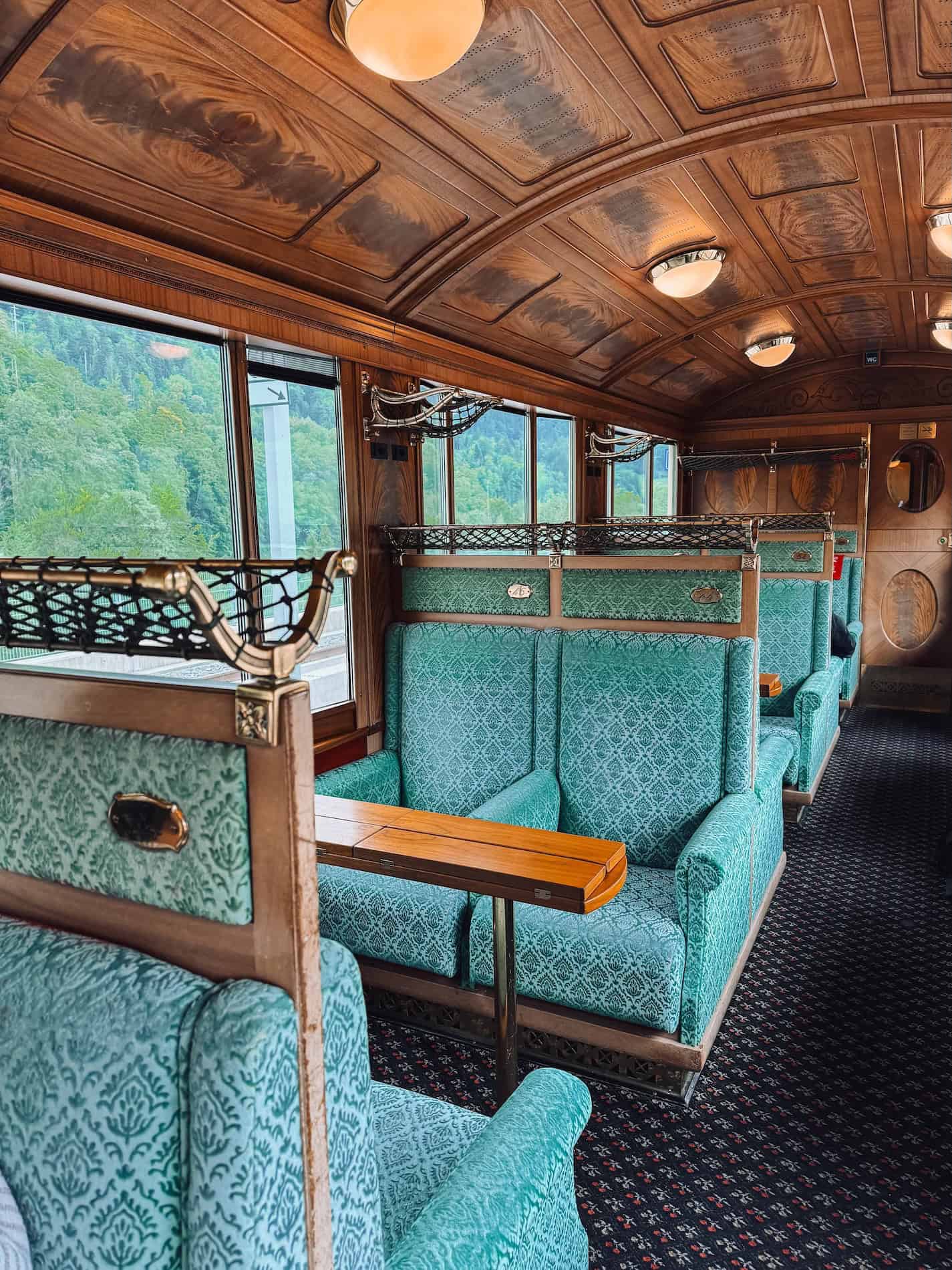 Side view inside the GoldenPass Belle Epoque train showing pairs of plush green upholstered seats, large panoramic windows overlooking Swiss mountain scenery, and beautifully detailed wooden interiors with brass luggage racks.