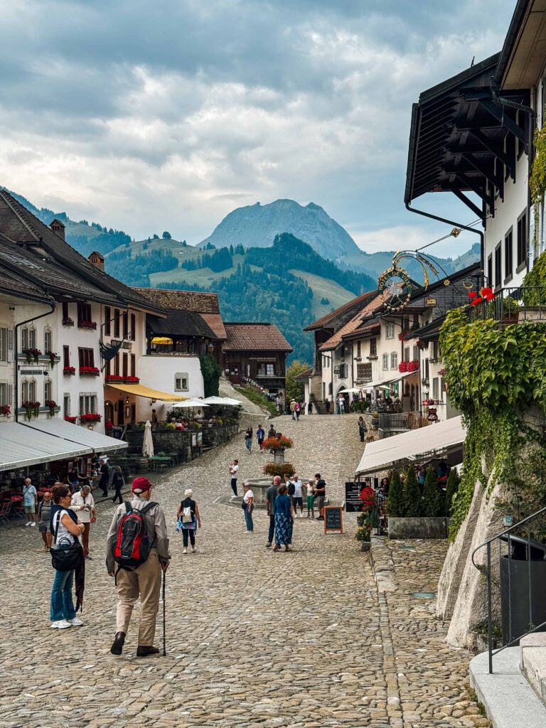 Cobblestone street in the medieval village of Gruyères, Switzerland, lined with charming buildings, restaurants, and mountain views in the distance.
