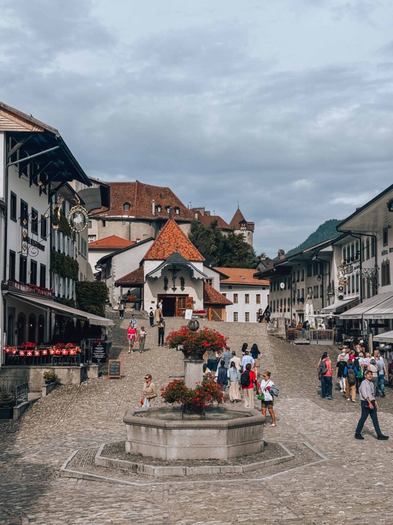 Central fountain surrounded by flowers and historic buildings in the old town of Gruyères, with visitors walking along the cobblestone street toward the castle. Gruyeres is a must visit in a 7 day Switzerland Itinerary. 