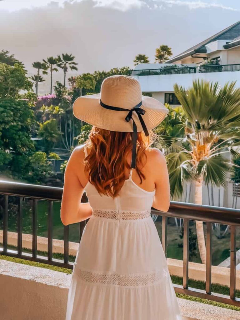 Woman wearing a white dress and wide-brimmed hat standing on a balcony surrounded by palm trees at a Maui resort during golden hour.
