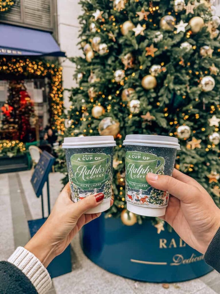 Two hands holding festive holiday cups of Ralph’s Coffee in front of a beautifully decorated Christmas tree with gold ornaments and twinkling lights outside the Ralph Lauren store.