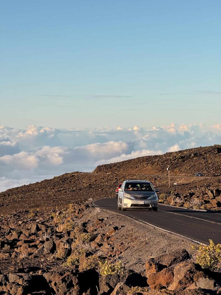 A car driving along the winding Crater Road near the summit of Haleakalā, surrounded by rocky volcanic terrain and clouds below.