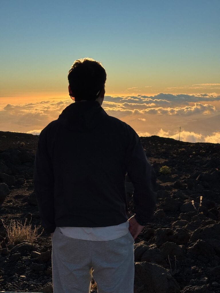 A man facing the colorful sunset on haleakalā over the clouds, standing on volcanic terrain under the warm evening light.