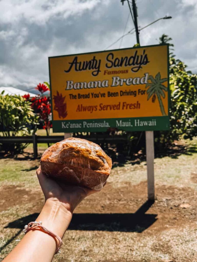 Hand holding a loaf of Aunty Sandy’s famous banana bread in front of the yellow roadside sign on Ke‘anae Peninsula, Maui