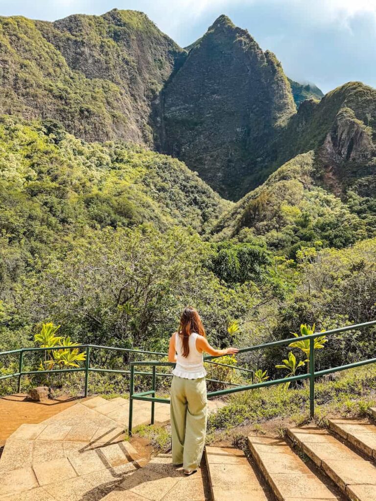 Woman standing on stone steps at a lookout point in Iao Valley State Park, admiring the towering emerald cliffs of the West Maui Mountains.