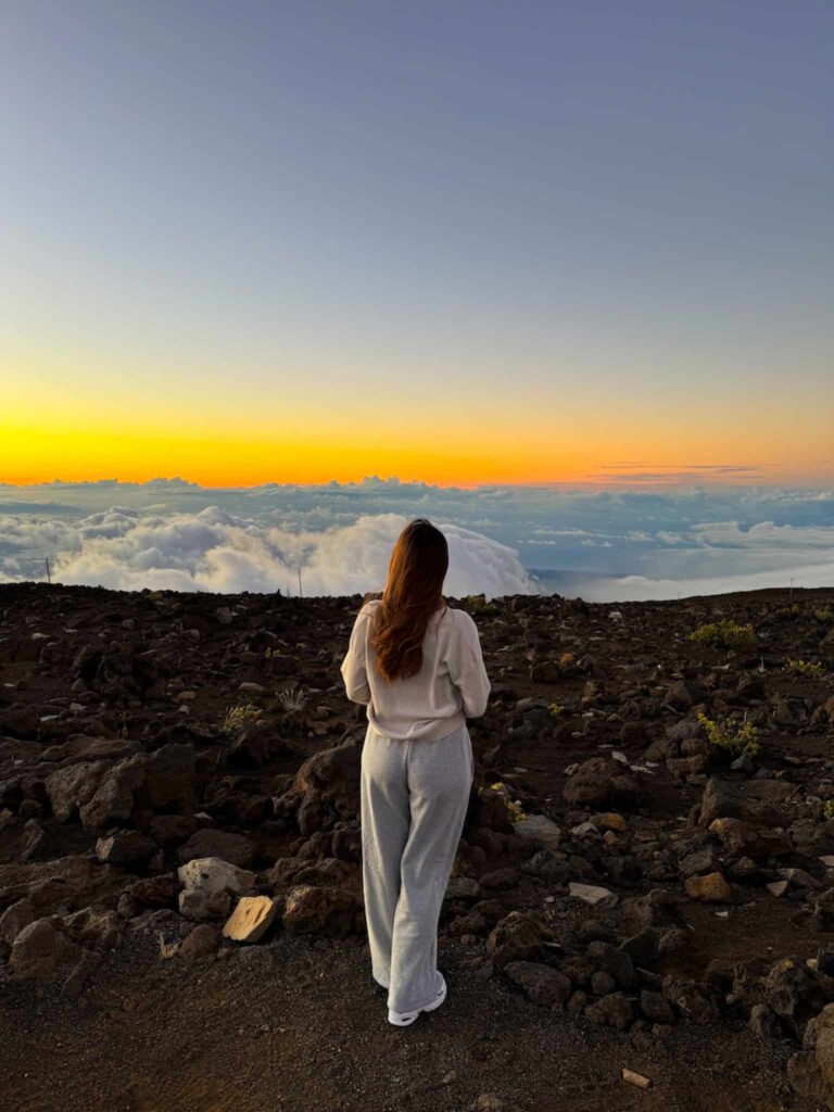 A woman standing on the road at Haleakalā Summit with arms open, enjoying the pastel sunset above the clouds.