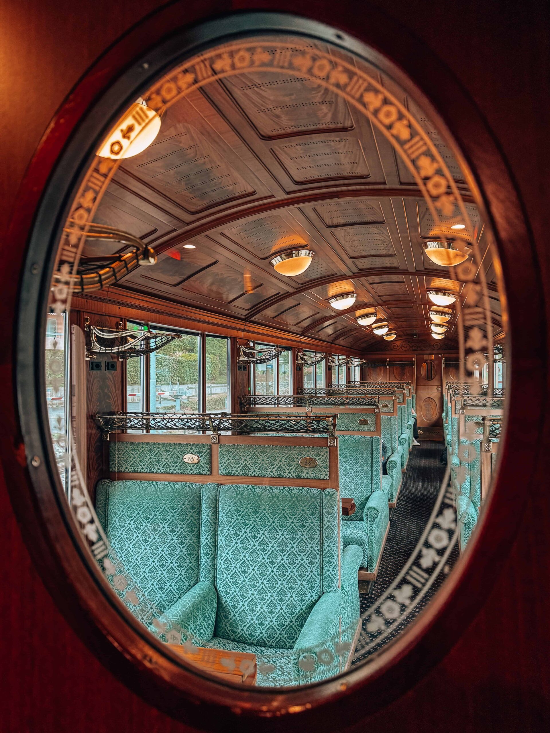 View through a circular glass window into the Belle Époque train carriage, revealing its ornate wooden ceiling, retro turquoise seats, and warm ambient lighting.