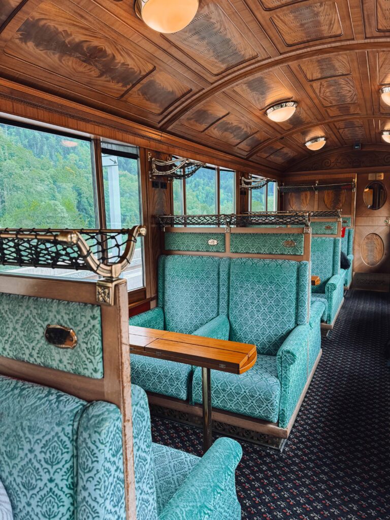 Elegant vintage train interior of the Belle Époque carriage with turquoise patterned seats, wooden paneling, and large windows overlooking the Swiss countryside.