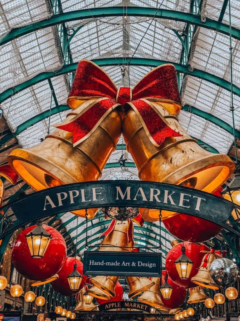 The Apple Market in Covent Garden beautifully decorated for Christmas with oversized golden bells, red bows, lanterns, and sparkling ornaments hanging from the glass ceiling.