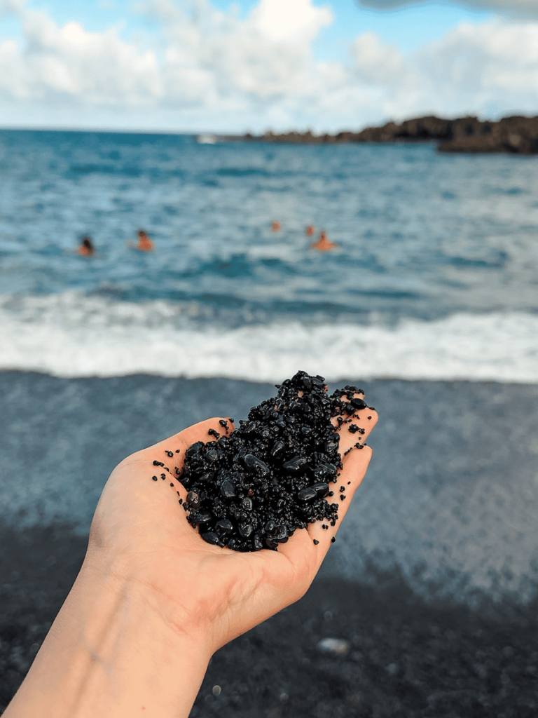 Close-up of a hand holding black volcanic sand at Waiʻanapanapa State Park in Maui, with swimmers enjoying the ocean in the background.