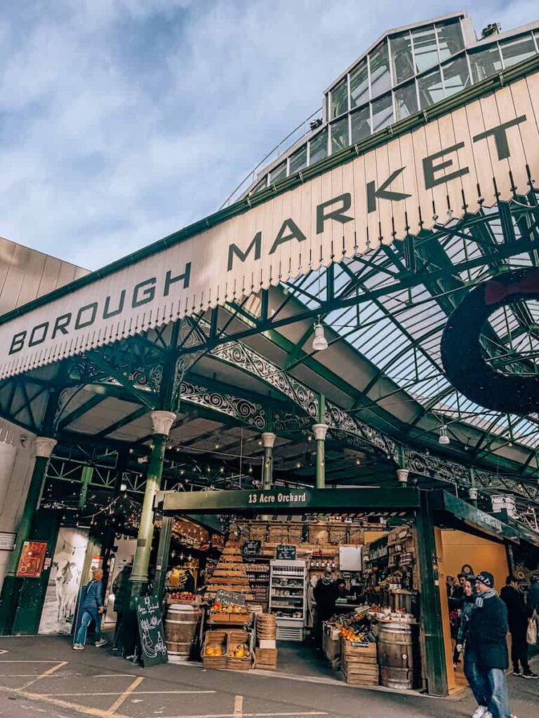 The main entrance of Borough Market in London with its iconic green iron framework and “Borough Market” sign, showing bustling stalls filled with fresh produce and artisanal goods.