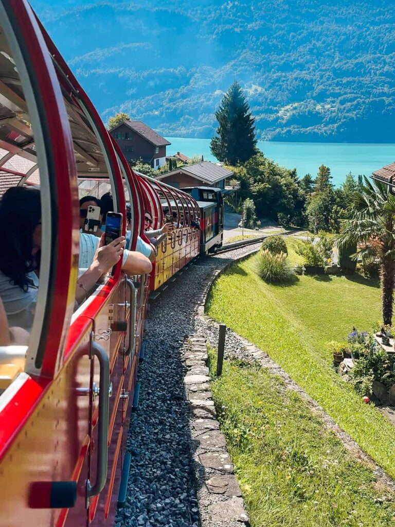 Passengers leaning out of the open-air red Brienz Rothorn Train as it winds past homes and turquoise Lake Brienz under clear blue skies.