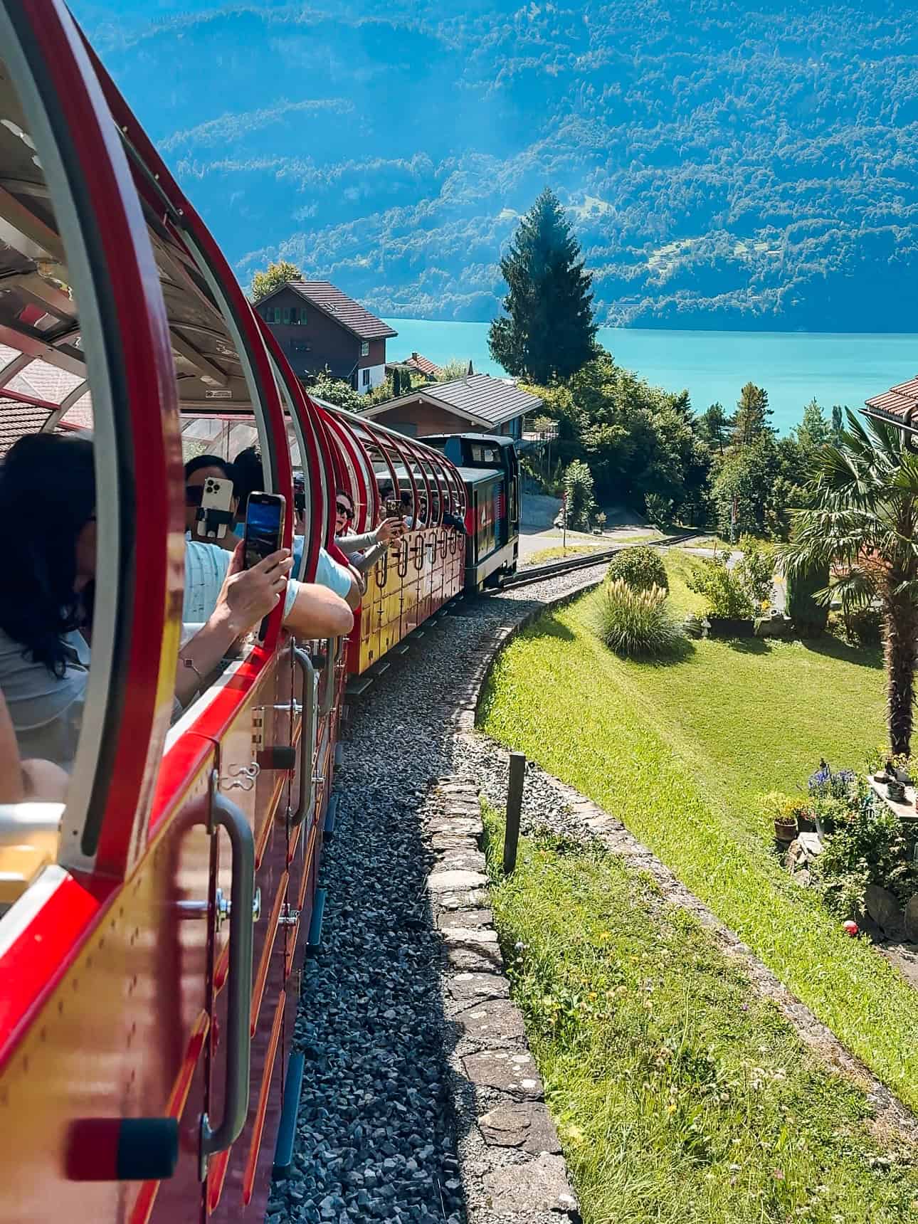 Passengers leaning out of the open-air red Brienz Rothorn Train as it winds past homes and turquoise Lake Brienz under clear blue skies.