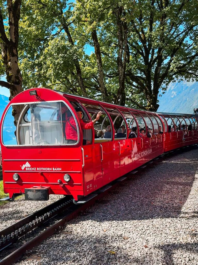 Bright red Brienz Rothorn Bahn train waiting at the station surrounded by tall green trees and mountain views in the background.