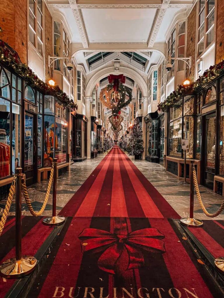 The Burlington Arcade in London beautifully decorated for the holidays with garlands, Christmas trees, and a luxurious red carpet featuring a ribbon design leading down the festive shopping corridor.