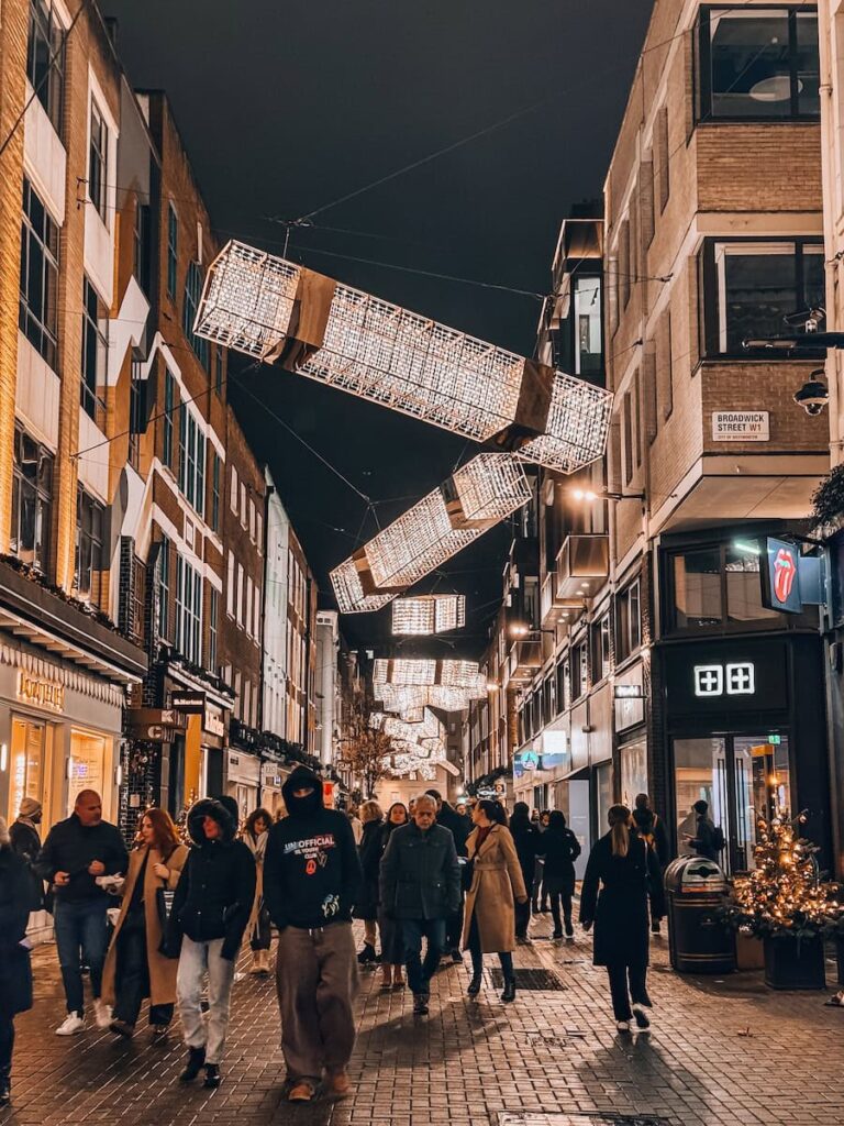 Crowds of people walking along Carnaby Street in London at night under creative illuminated cube-shaped Christmas lights, surrounded by festive shopfronts and warm holiday decor.