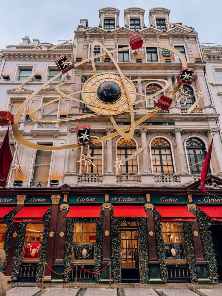 The Cartier store in London adorned with festive Christmas decorations, including a grand gold clock display with orbiting red gift boxes and stars above the elegant red-and-green storefront.