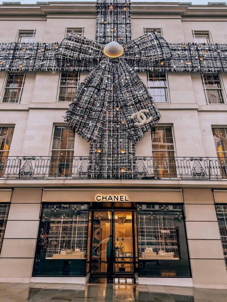 The exterior of the Chanel boutique in London decorated for Christmas with a massive black and gold bow made of tweed fabric draped across the building facade, glowing lights accenting the elegant holiday display.