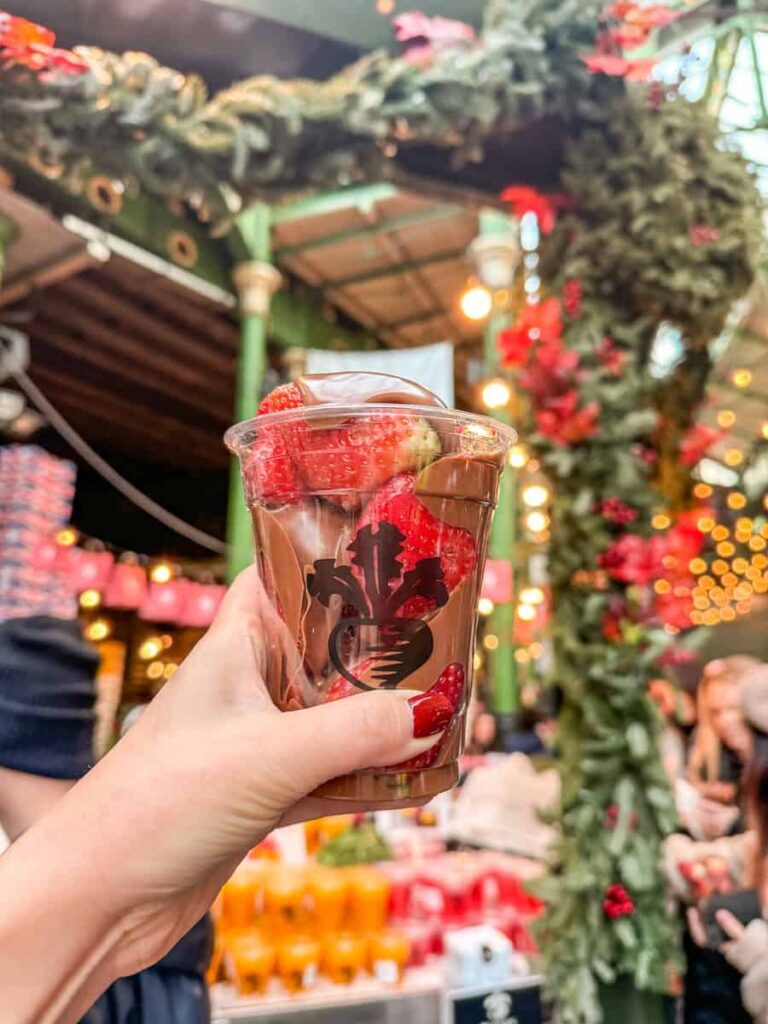 A hand holding a cup of fresh strawberries topped with rich melted chocolate inside Borough Market, with festive holiday decorations and lights in the background.