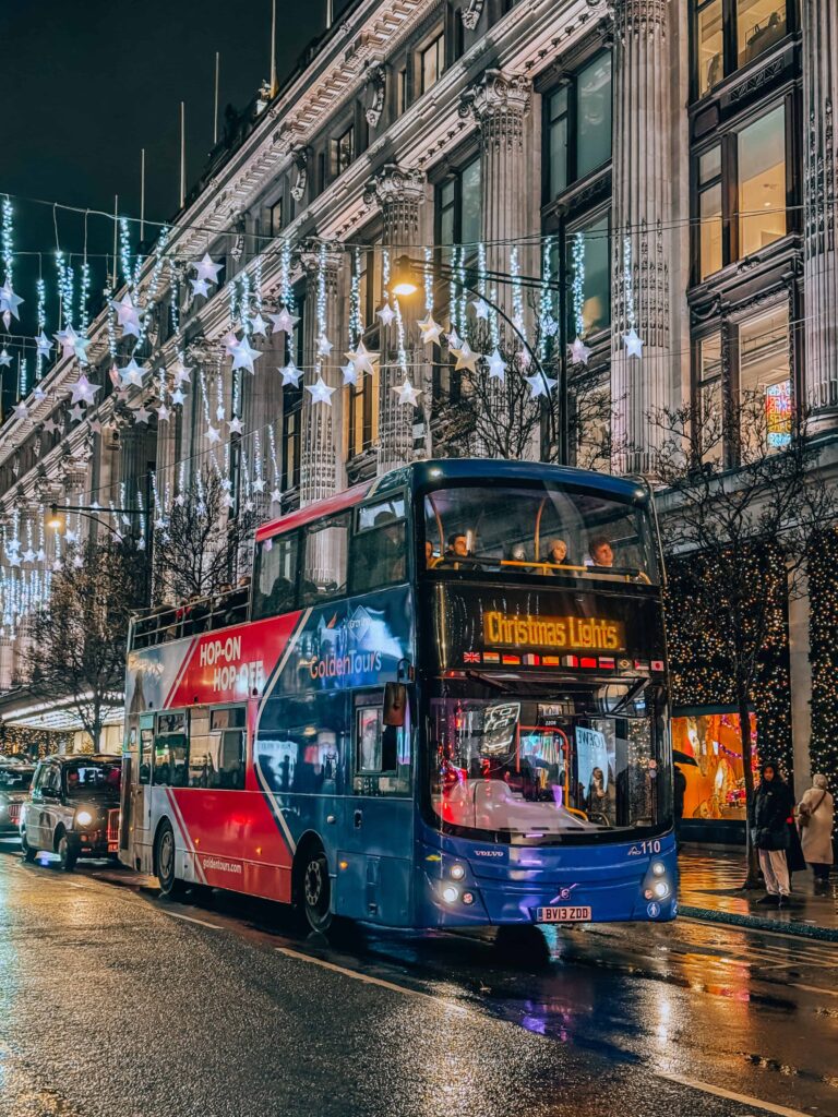 A festive double-decker “Hop-On Hop-Off” Christmas Lights tour bus driving through central London at night, surrounded by glowing star-shaped lights and holiday decorations along the street.