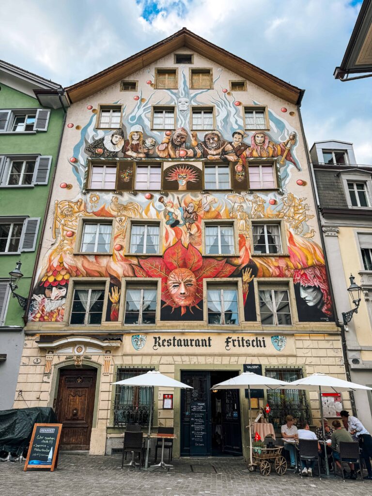 Colorful mural-covered facade of Restaurant Fritschi in Lucerne, Switzerland, depicting playful figures and vibrant scenes related to Fasnacht festival, with outdoor tables and umbrellas on the cobblestone street.