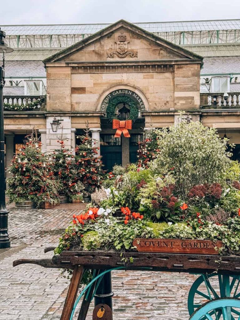 The exterior of Covent Garden Market adorned with holiday decorations, including Christmas trees and a large wreath with a red bow, with a rustic flower cart labeled “Covent Garden” filled with colorful greenery and blooms in the foreground.