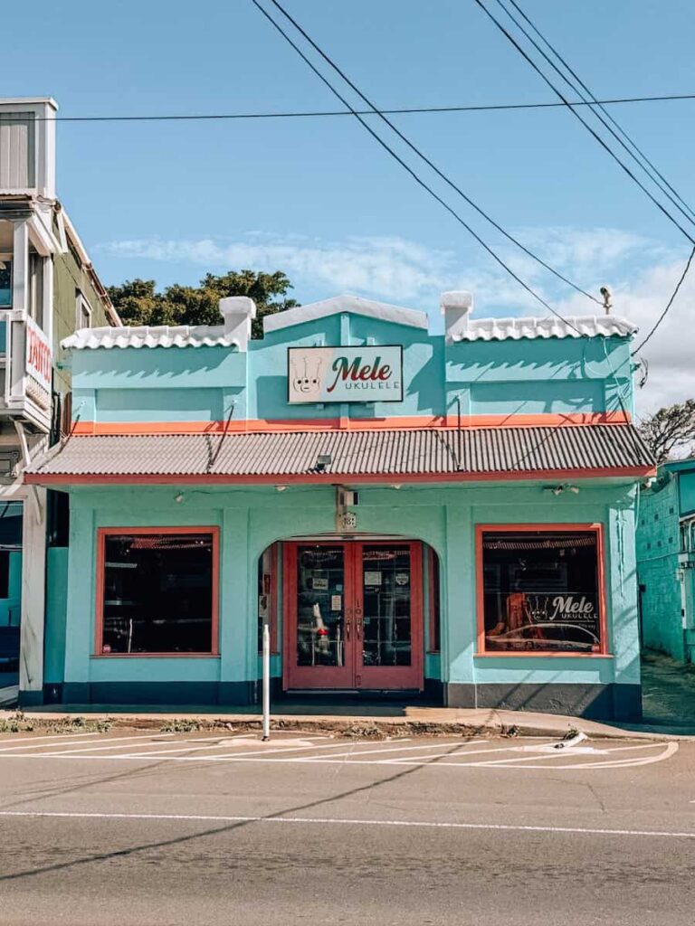 Colorful turquoise storefront of Mele Ukulele shop in Paia, Maui, with coral-red trim and a bright blue sky overhead.