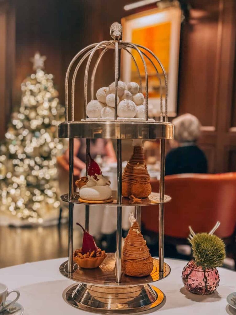 A festive afternoon tea display with desserts arranged on a two-tiered silver stand shaped like a birdcage, featuring pear-shaped pastries and snowball sweets, with a Christmas tree softly glowing in the background at Carlton Tower Jumeirah in London during Christmas. 