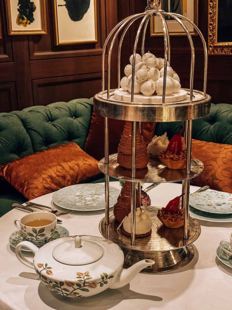 An elegant afternoon tea setup during Christmas in London with a floral teapot and teacup, accompanied by a tiered dessert stand shaped like a birdcage filled with pastries and sweets, set against a cozy, warmly lit interior.