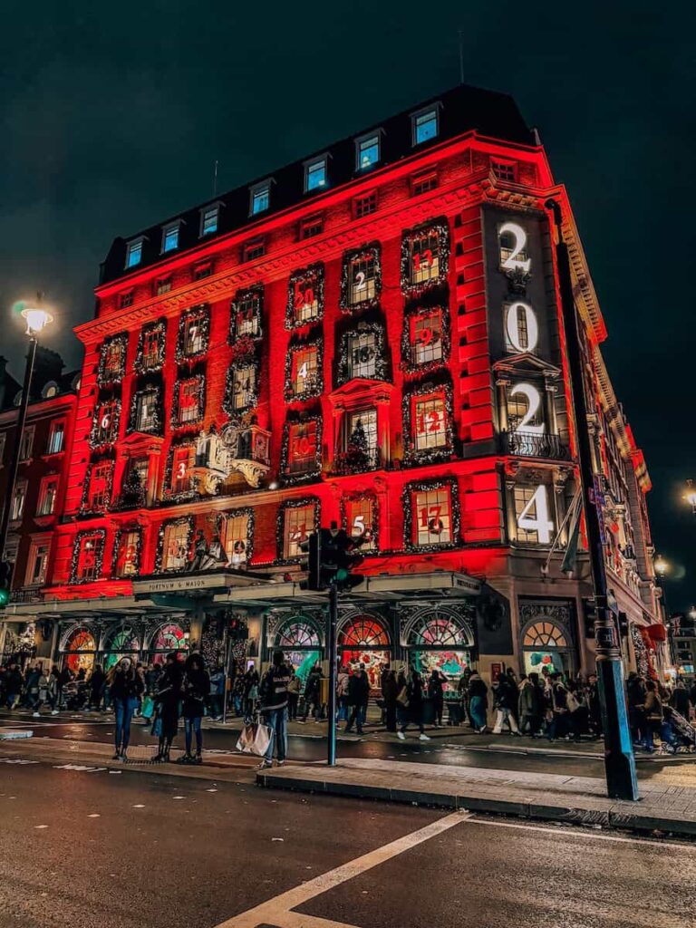 The Fortnum & Mason building in London glowing red at night, transformed into a giant advent calendar with numbered windows and festive garlands, as shoppers gather outside during the holiday season.