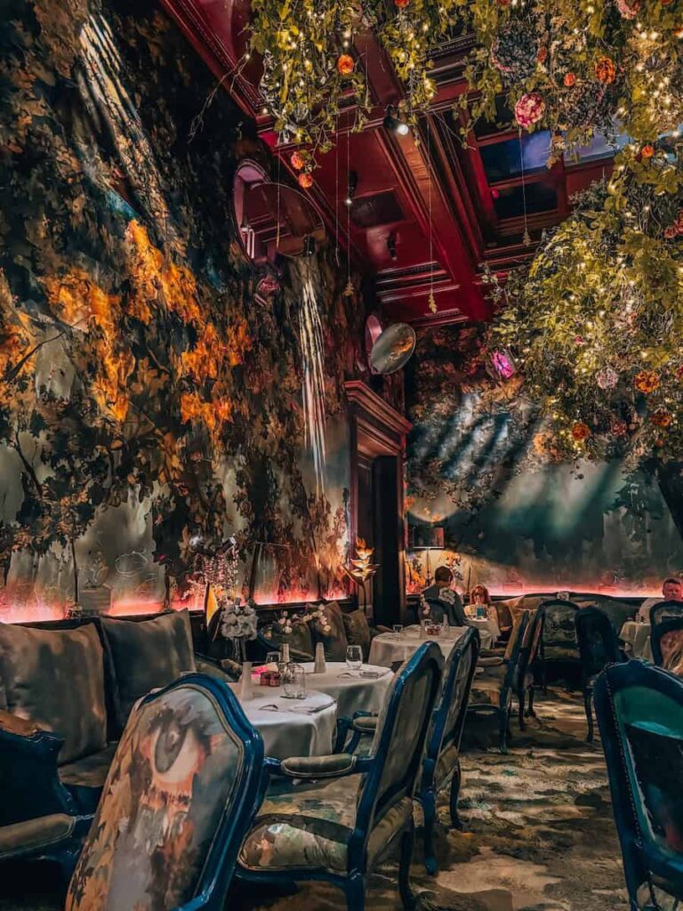A dimly lit, romantic restaurant interior with floral ceiling installations, lush greenery, and dramatic mural-covered walls, creating an enchanting forest-like atmosphere.