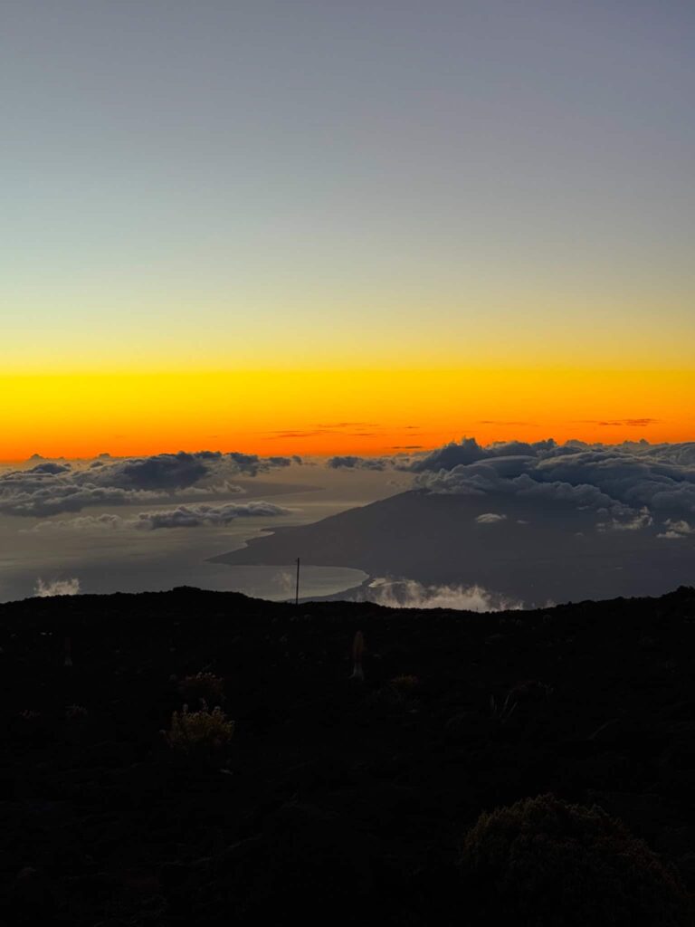 The sun setting over layers of clouds at Haleakalā National Park, painting the sky in deep orange and pink hues as the light fades.