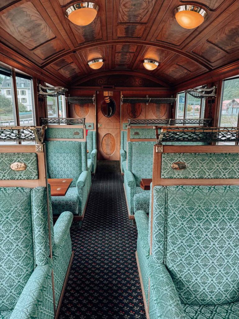 Interior of the GoldenPass Belle Epoque train first-class carriage featuring elegant green velvet seats, polished wood paneling, brass details, and warm ceiling lights that create a vintage luxury ambiance