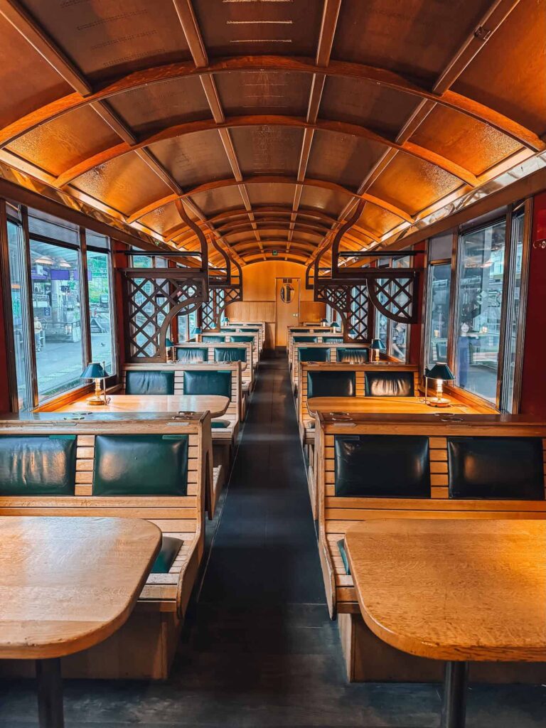 Interior of the GoldenPass Belle Epoque train second-class carriage with wooden booth-style seating, dark green leather cushions, warm lighting, and a curved wood-paneled ceiling that evokes a cozy vintage café atmosphere.