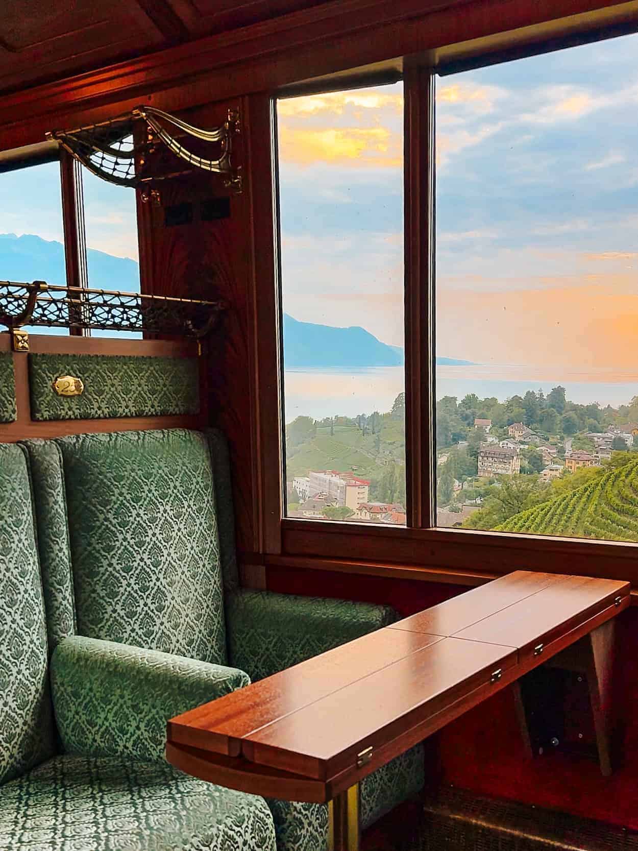 A plush green velvet seat and polished wooden table inside the GoldenPass Belle Epoque train, with large windows framing a stunning view of vineyards, Lake Geneva, and distant mountains under a pastel-colored sunset sky.