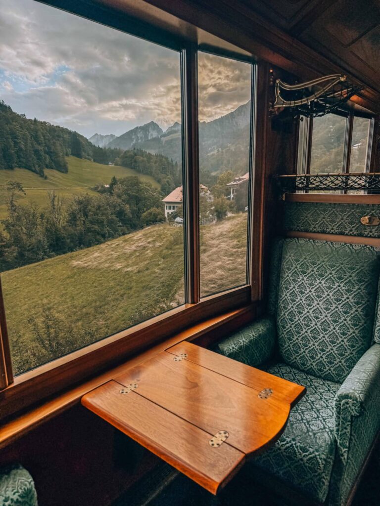 A single green velvet seat and wooden fold-out table inside the GoldenPass Belle Epoque train, with large panoramic windows revealing a serene view of rolling hills, alpine houses, and distant mountains under a moody sky.