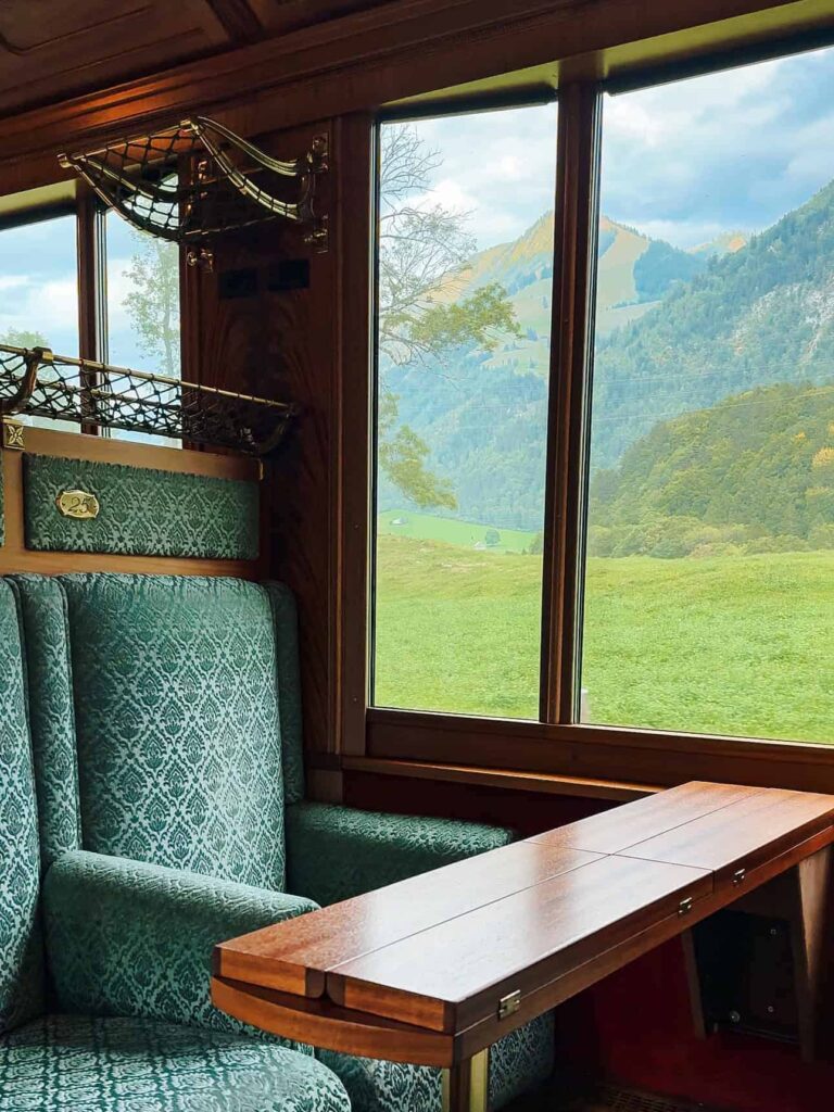Alt text: A plush green velvet seat and wooden table inside the GoldenPass Belle Epoque train with large panoramic windows framing a view of rolling green hills and mountains under a cloudy sky.