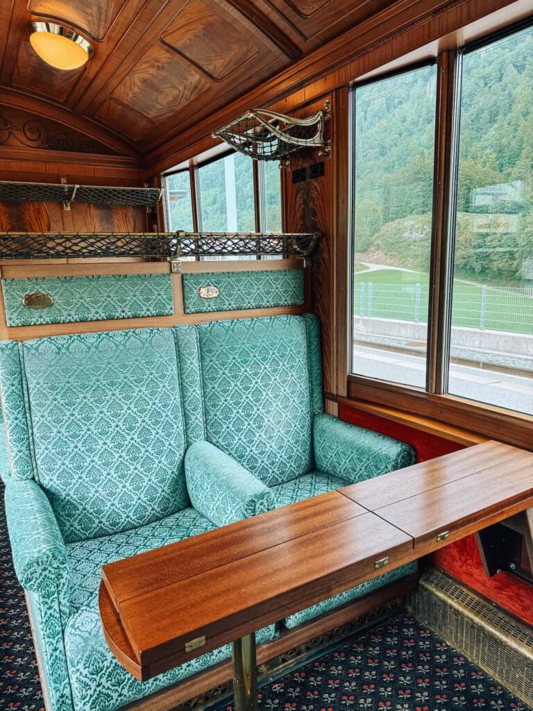 Elegant green velvet seats with a wooden table inside the GoldenPass Belle Epoque train, featuring vintage wood paneling, brass luggage racks, and large panoramic windows overlooking the Swiss countryside.